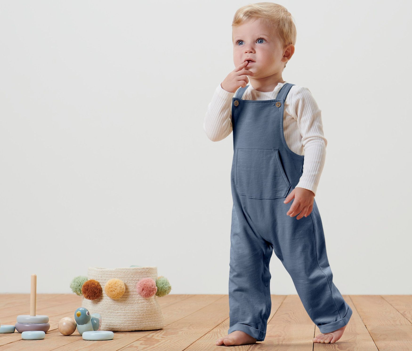 Ein kleines Kind steht auf einem Holzboden und trägt eine blaue Baby-Latzhose.