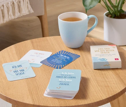 Auf einem Holztisch stehen eine hellblaue Tasse mit einem Getränk und ein Gelassenheits-Karten-Set »Lass Dich von den Wellen tragen«.