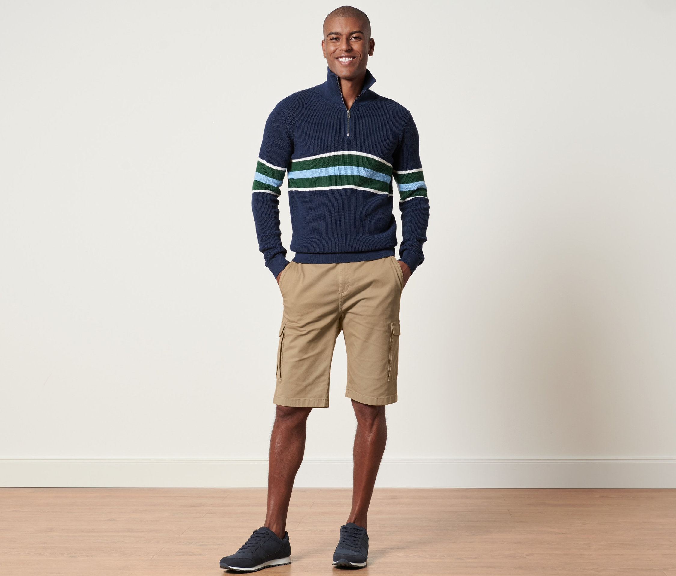 Mann steht und posiert in einem marineblauen Streifenpullover und sandfarbenen Cargo-Shorts.