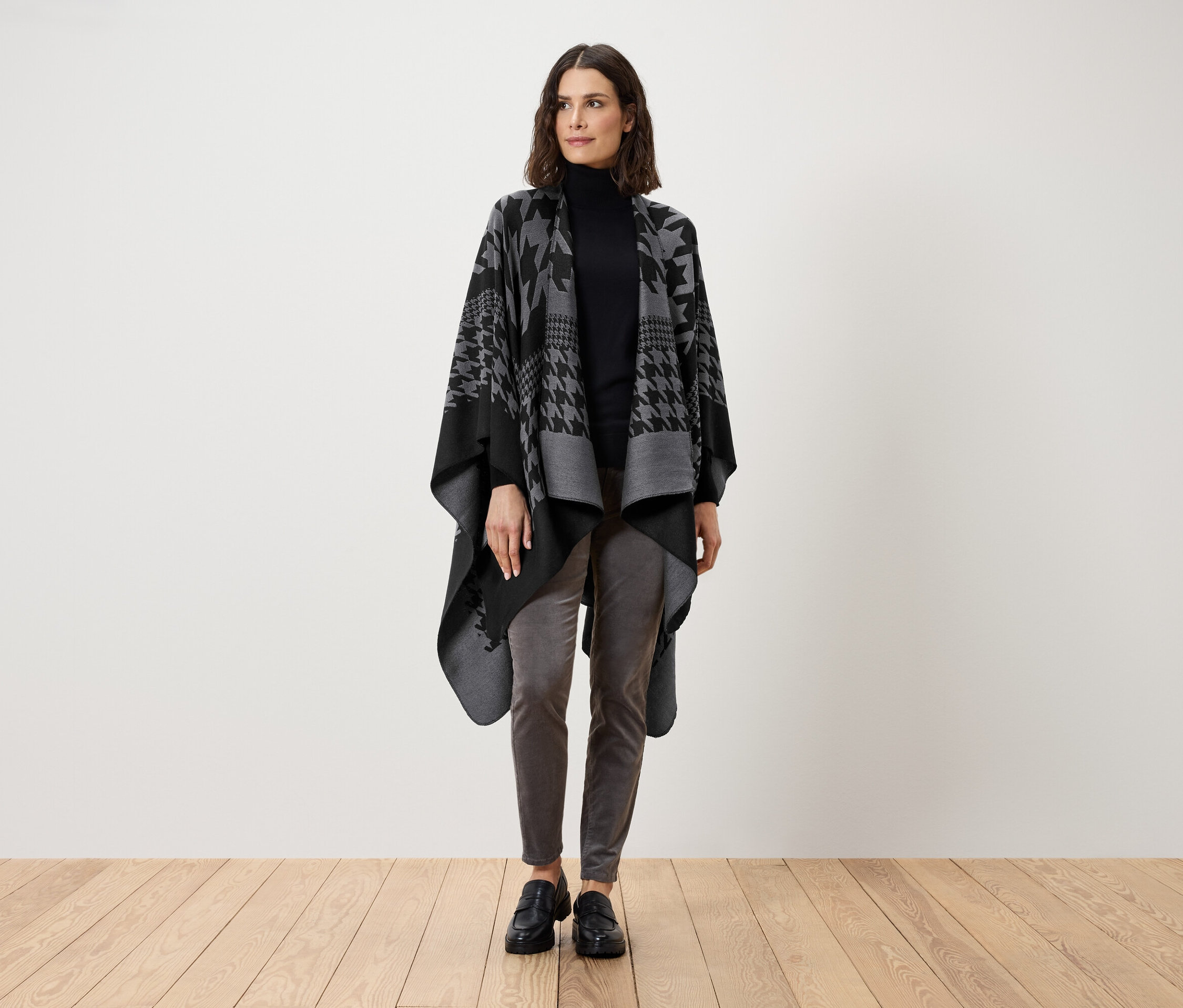 Eine Frau steht und trägt einen schwarzen Rollkragenpullover, graue Hosen und eine grau-schwarze Cape mit Hahnentrittmuster.