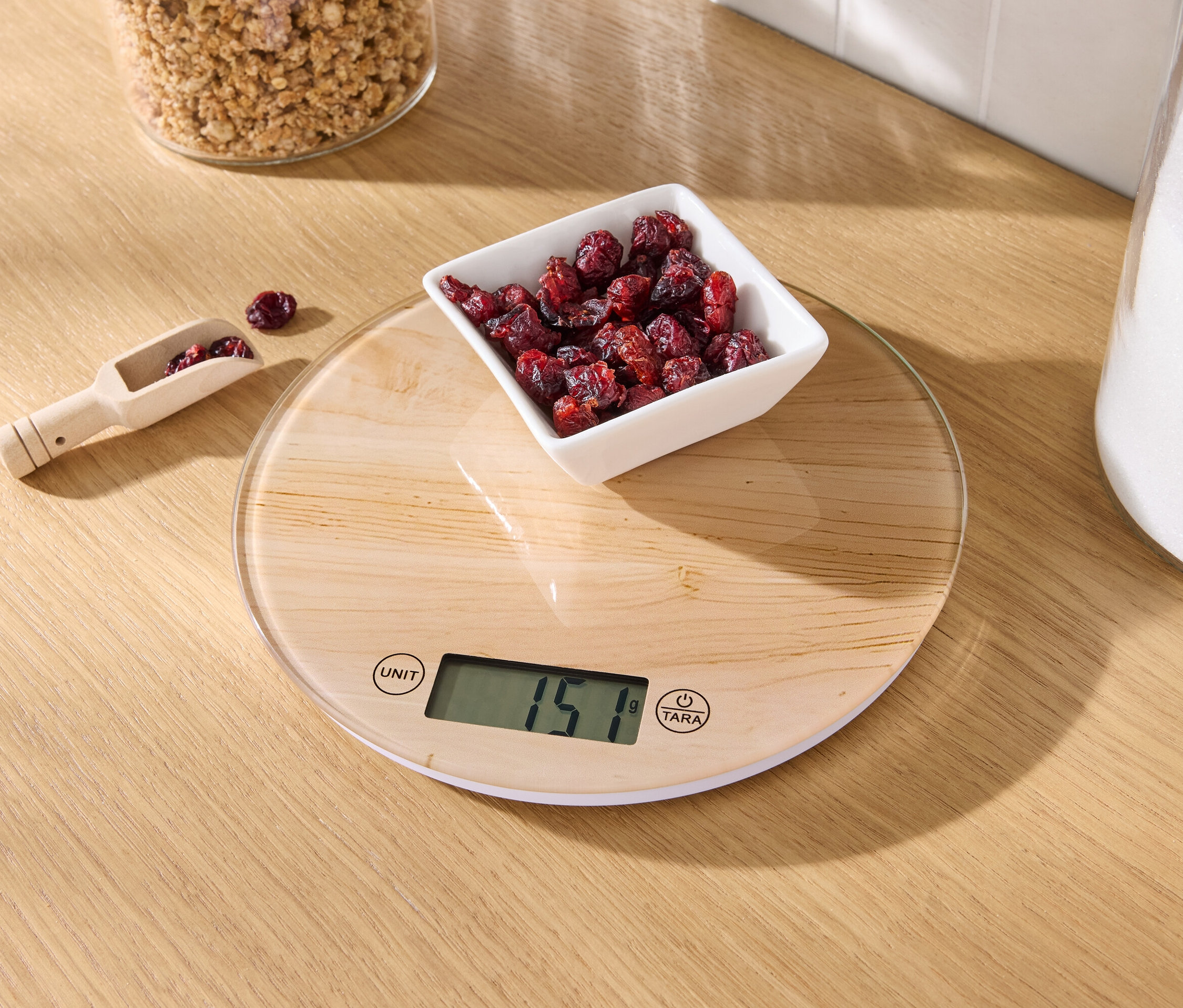 Auf einem Holztisch steht eine Küchenwaage mit einer Schale getrockneter Cranberries und einem hölzernen Messlöffel mit Cranberries.