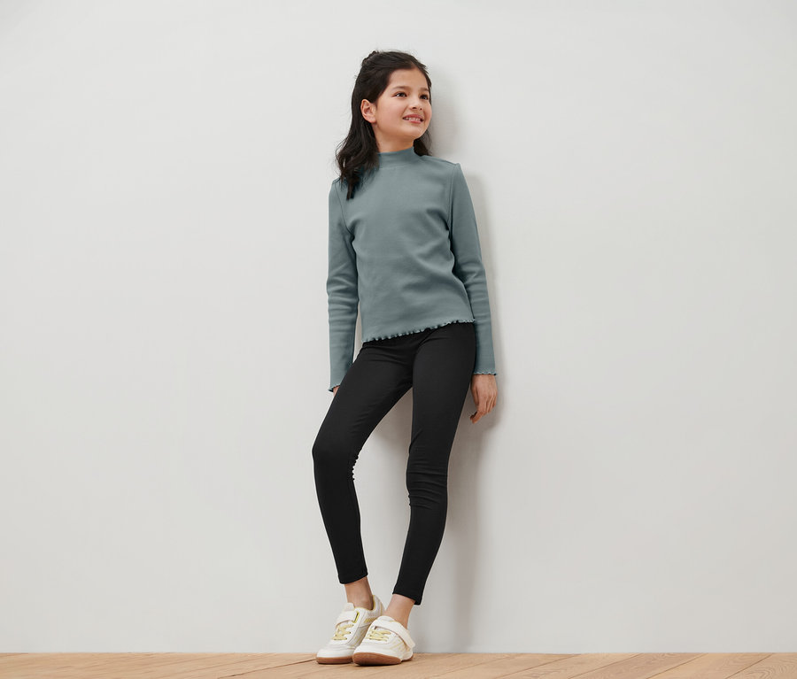 Lächelndes junges Mädchen in einem blaugrünen Langarmshirt, schwarzen Jeans und weißen Turnschuhen posiert vor einer weißen Wand.