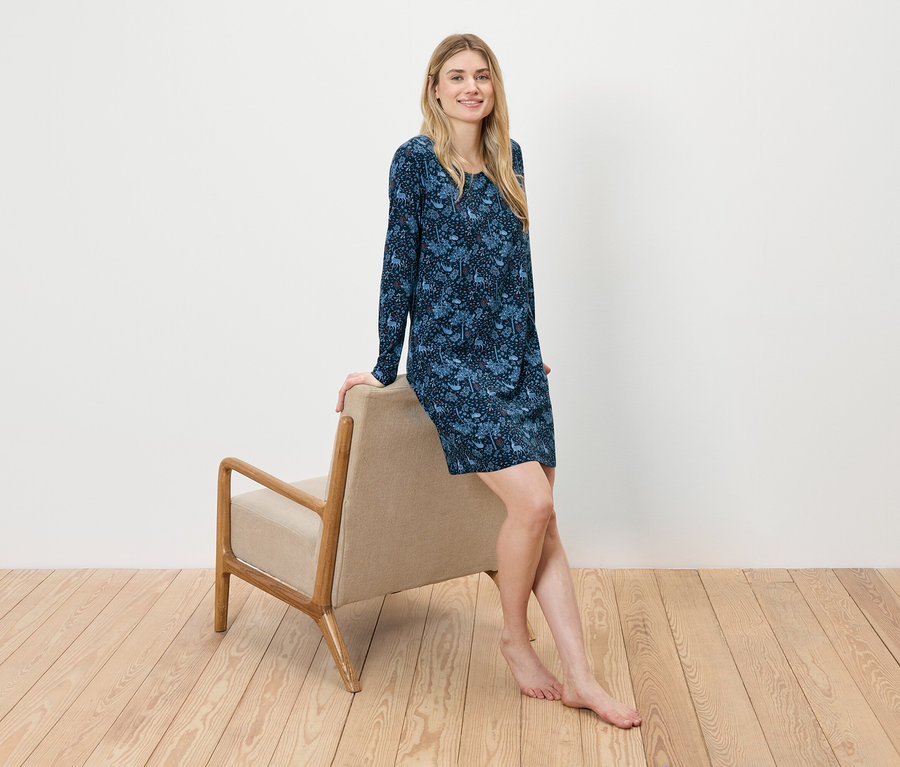 Eine Frau sitzt an einem beigen Sessel gelehnt und trägt ein blaues Blumenkleid.