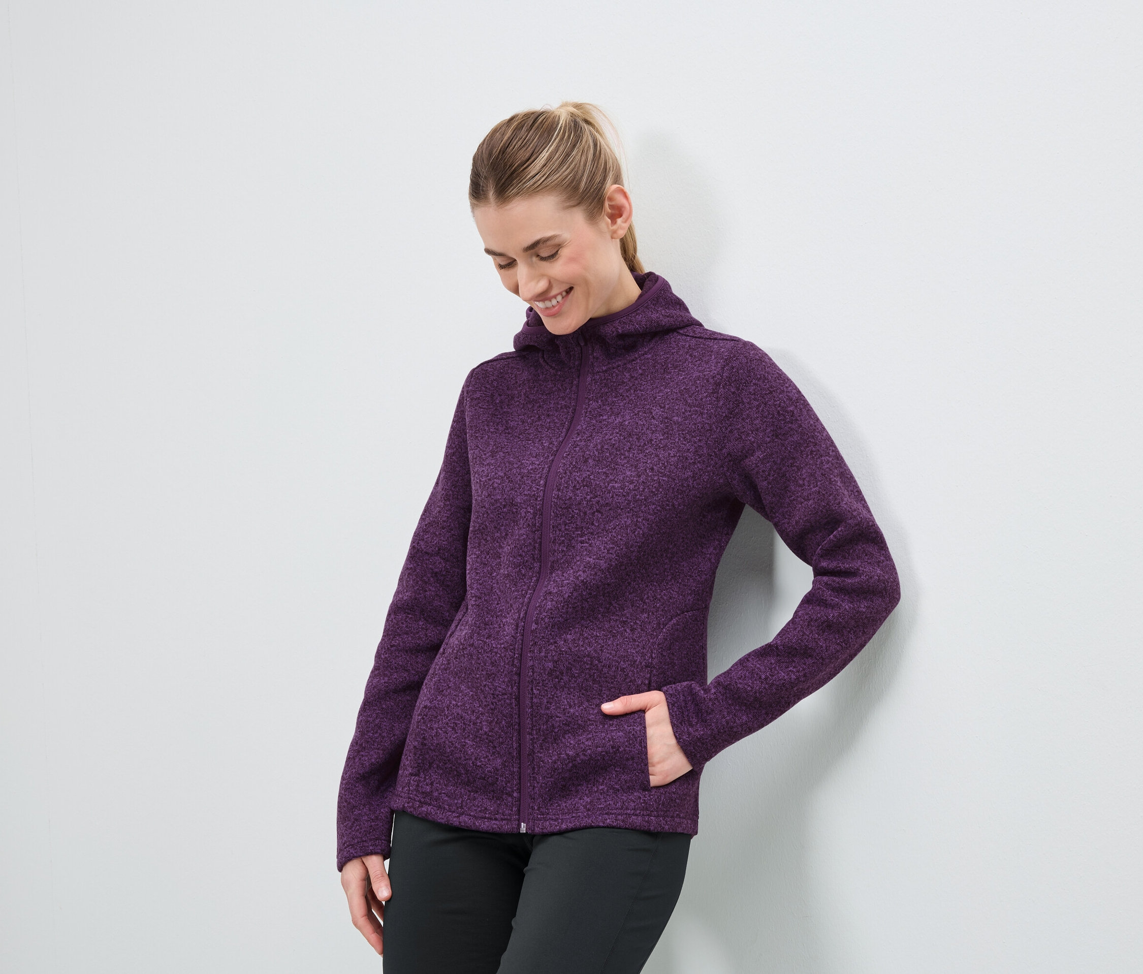 Eine Frau in einer lila Kapuzenjacke mit Reißverschluss und einer schwarzen Hose steht an einer weißen Wand.