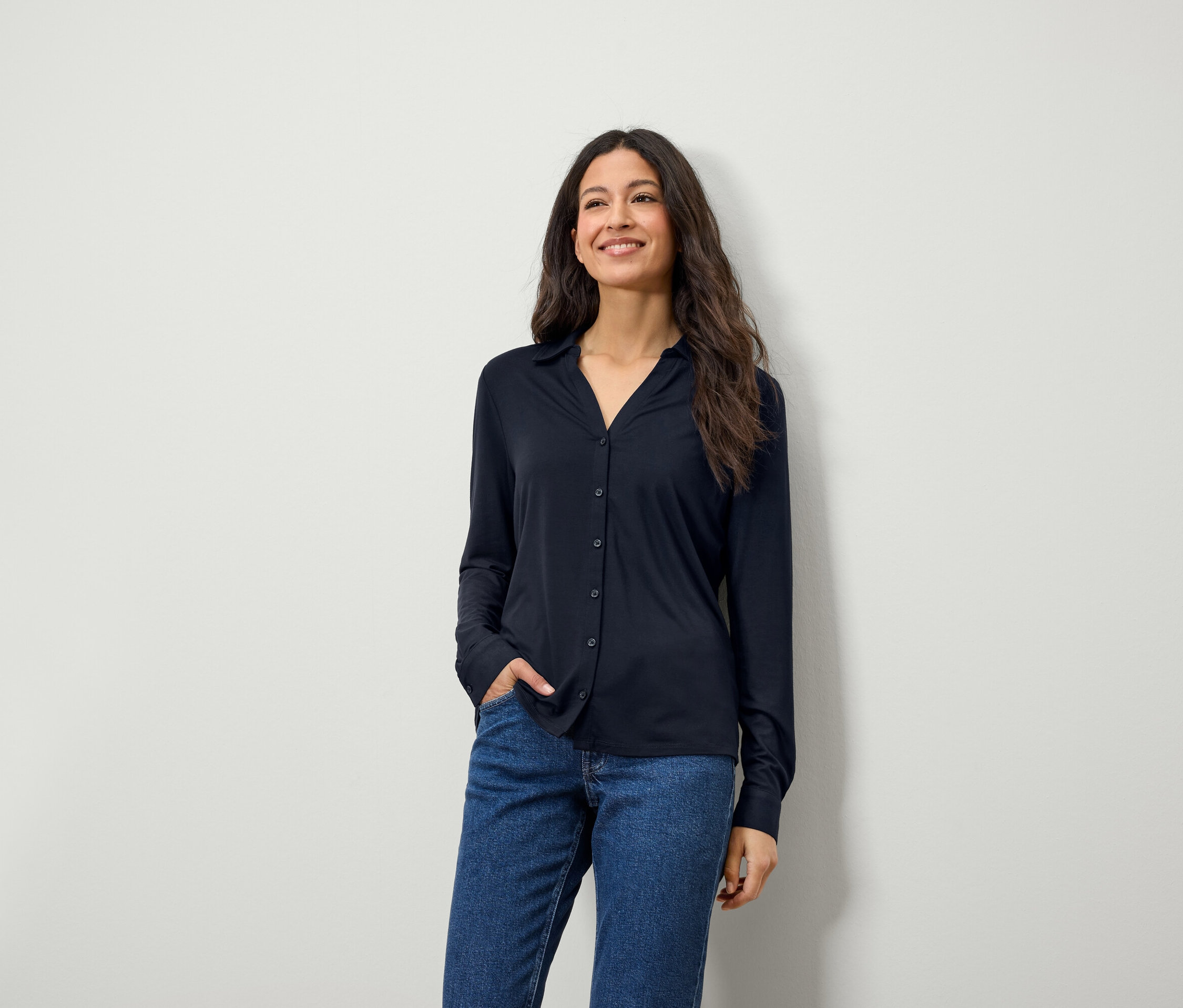 Eine lächelnde Frau mit langen braunen Haaren posiert in einem blauen Langarmhemd und Jeans.