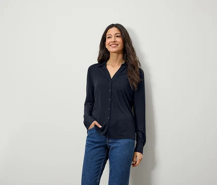 Eine lächelnde Frau mit langen braunen Haaren posiert in einem blauen Langarmhemd und Jeans.