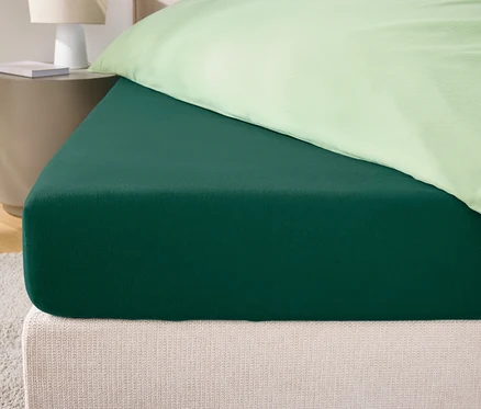 Detailaufnahme eines grünen Spannbettlakens auf einem Bett mit cremefarbenem Kopfteil und hellgrünem Kissenbezug.