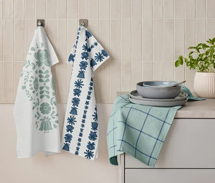 Zwei Geschirrtücher mit floralen Mustern hängen nebeneinander an der Wand. Darunter liegt ein gefaltetes grünes Geschirrtuch mit blauen Streifen, auf dem Teller und eine Schüssel gestapelt sind. Daneben steht ein Topf mit Kräutern.