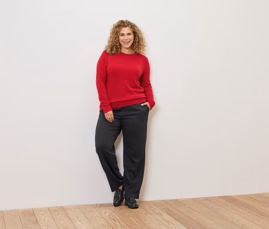Eine Frau mit lockigem Haar lehnt an einer Wand und trägt einen roten Pullover und eine dunkle Hose.