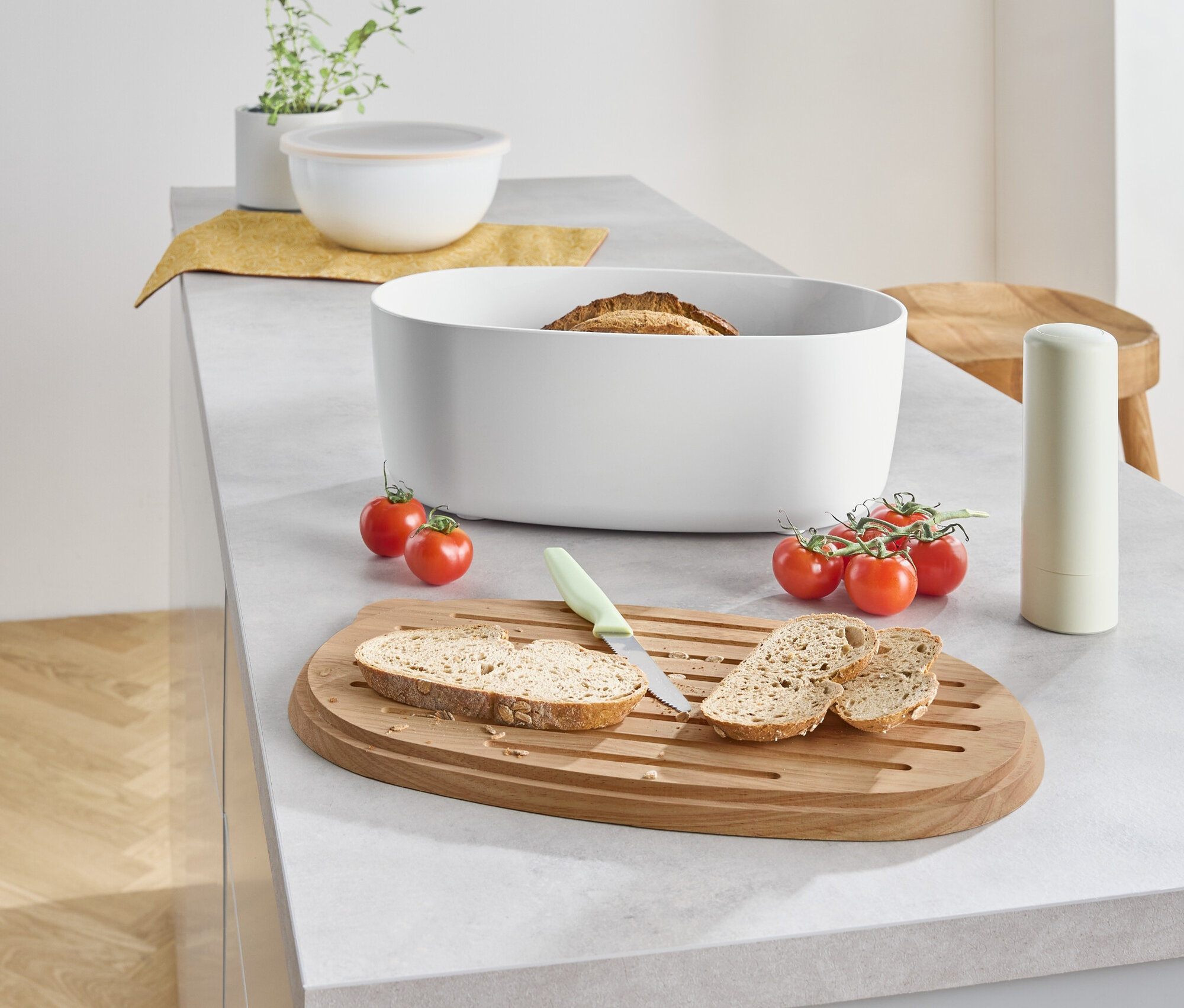 Auf einer Küchentheke liegen Brot in einem weißen Behälter, daneben geschnittenes Brot auf einem Holzbrett mit einem Messer und Cherrytomaten.