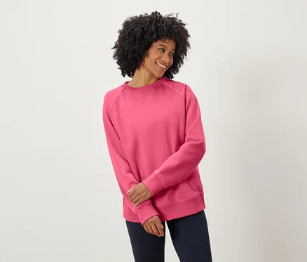 Frau mit dunklem lockigem Haar lächelt und trägt ein rosa Sweatshirt und marineblaue Hosen.