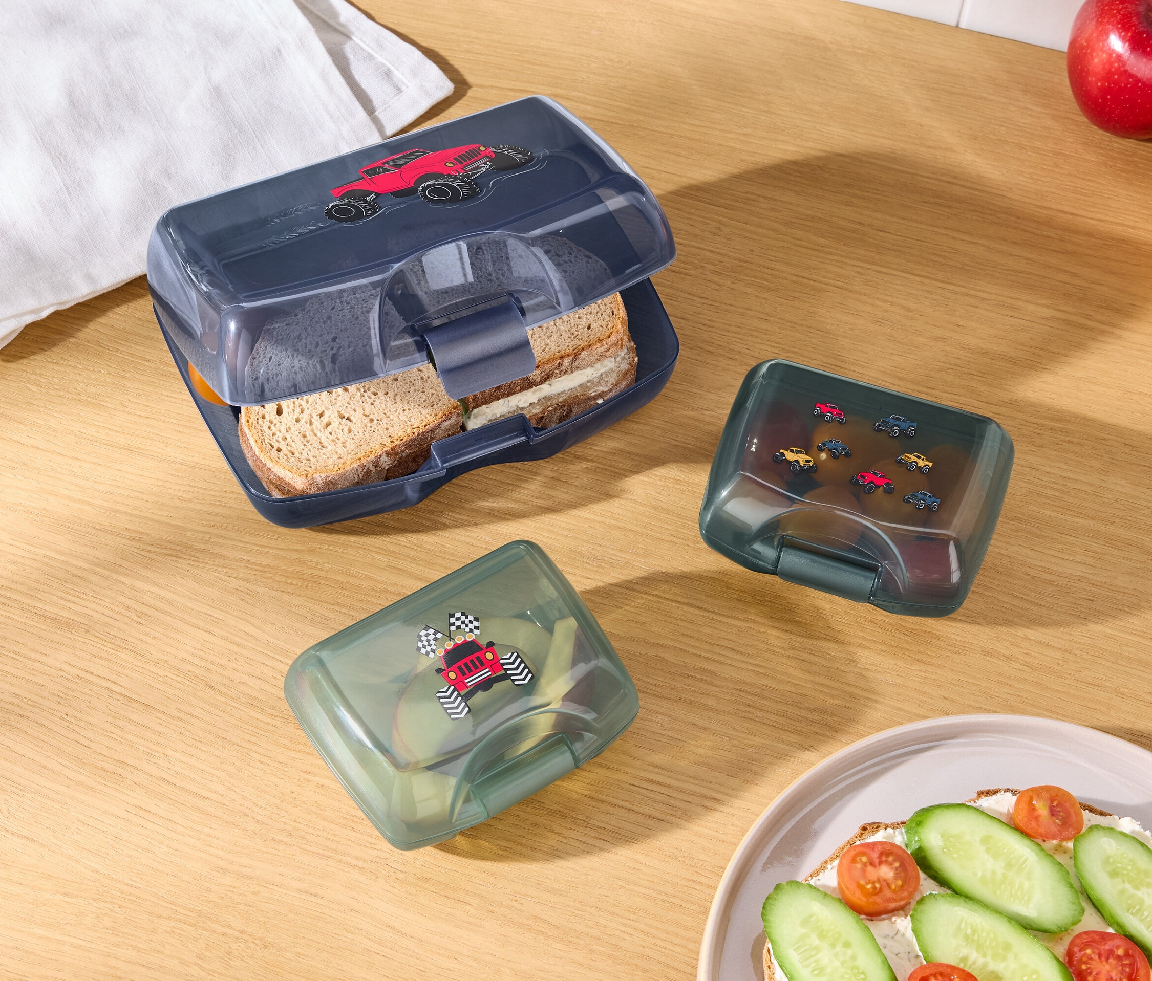 Auf einem Holztisch liegen eine geöffnete blaue Brotdose mit einem roten Auto darauf, zwei grüne Brotdosen mit Autoabbildungen, ein Apfel, eine Orange, ein weißes Tuch und ein Teller mit einem belegten Brot.