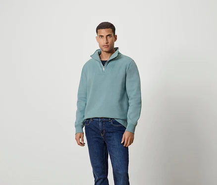 Mann steht und trägt ein blaugrünes Sweatshirt mit Reißverschluss und Jeans.