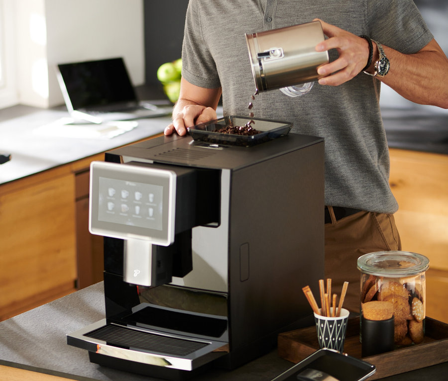 Mann mit Brille gießt Kaffeebohnen in den »Tchibo Office« Kaffeevollautomat, schwarz.