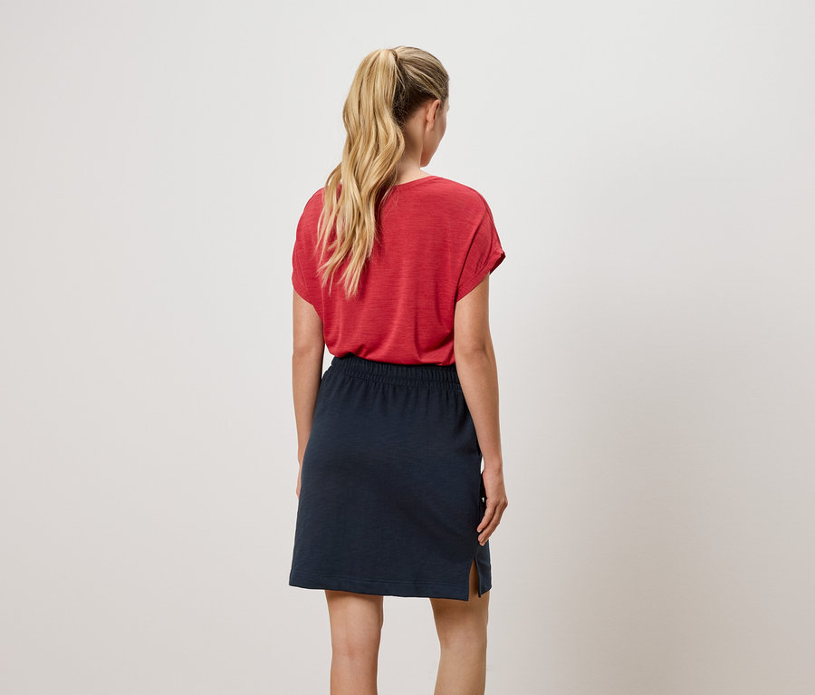 Rückansicht einer Frau mit blondem Pferdeschwanz, die ein rotes T-Shirt und einen marineblauen Rock trägt.