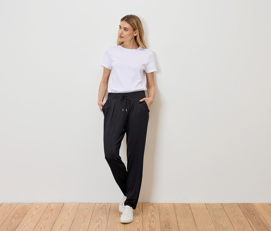 Frau steht mit Händen in den Taschen vor einer Wand, trägt ein weißes T-Shirt, eine schwarze Hose mit Kordelzug und weiße Turnschuhe.