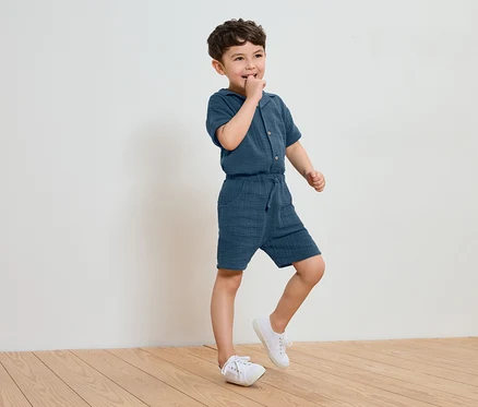 Ein kleiner Junge in einem blauen Baumwoll-Jumpsuit und weißen Turnschuhen steht auf einem Holzboden und lutscht an seinem Finger.