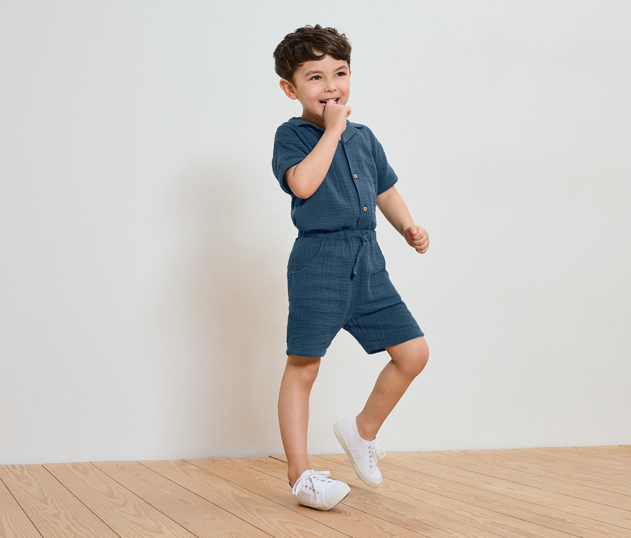 Ein kleiner Junge in einem blauen Baumwoll-Jumpsuit und weißen Turnschuhen steht auf einem Holzboden und lutscht an seinem Finger.