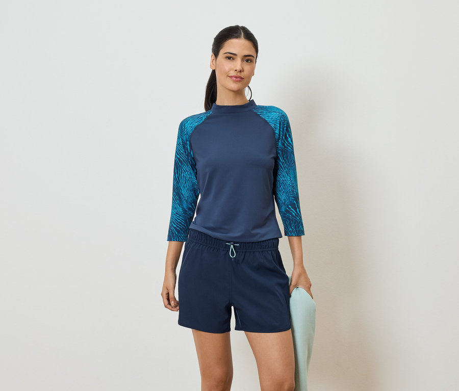 Frau mit marineblauem Oberteil mit Dreiviertelärmeln und blauen Shorts, hält ein hellblaues Handtuch vor einem weißen Hintergrund.