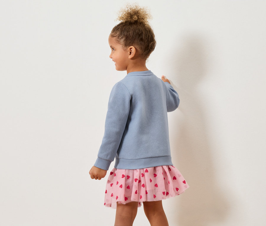 Mädchen von hinten in blauem Pullover und rosa Rock mit Herzen.