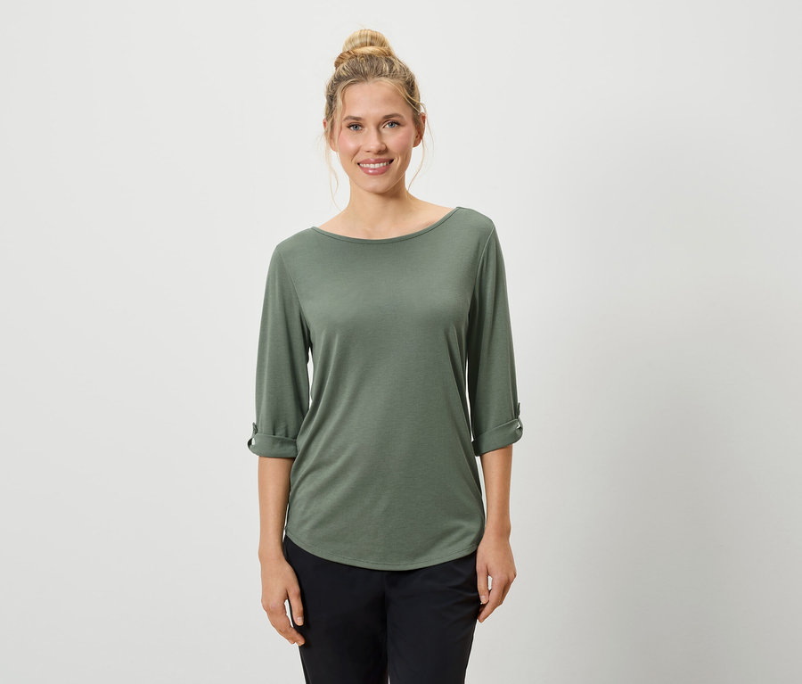 Eine Frau mit hochgesteckten blonden Haaren trägt ein olivgrünes Langarmshirt und schwarze Hosen vor einem weißen Hintergrund.