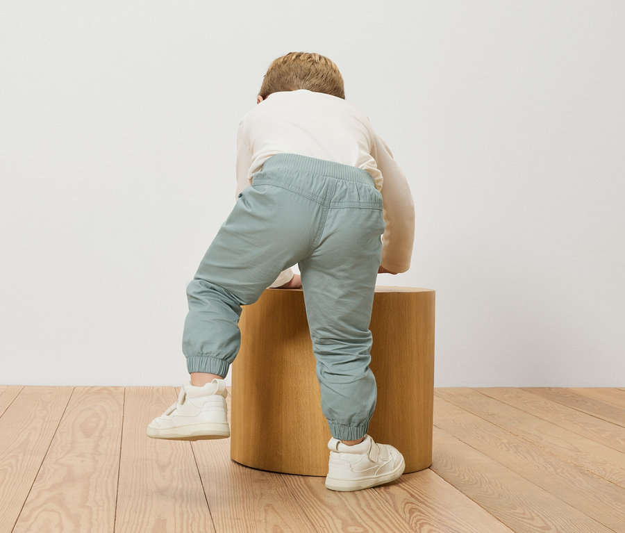 Ein Kind klettert auf einen hölzernen Hocker, trägt eine lange Ärmel und hellblaue Hosen.