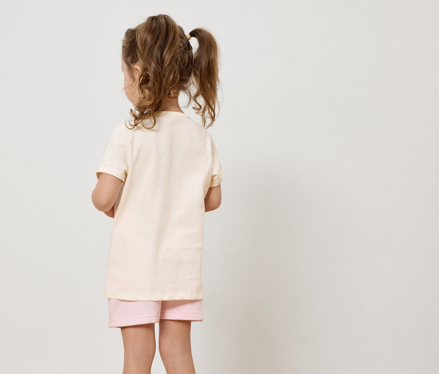 Ein Mädchen mit blonden Haaren, das ein beiges T-Shirt und rosa Shorts trägt, steht mit dem Rücken zur Kamera vor einem weißen Hintergrund.