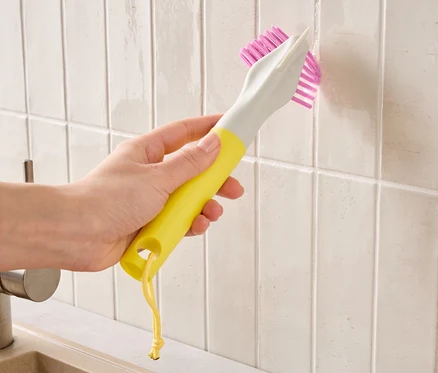 Eine Hand hält eine gelb-graue Bürste mit pinken Borsten und reinigt Fliesenfugen.
