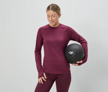 Frau in bordeauxfarbenem Sportoutfit hält einen 10 kg schweren Medizinball vor einem grauen Hintergrund.