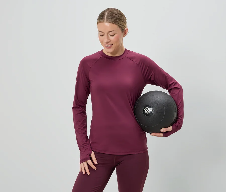 Frau in bordeauxfarbenem Sportoutfit hält einen 10 kg schweren Medizinball vor einem grauen Hintergrund.