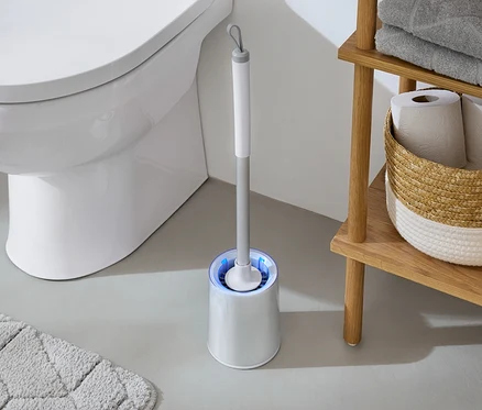 Im Badezimmer steht eine weiße Toilette neben einer WC-Bürste mit blauem Licht und einem Holzregal mit Handtüchern und Toilettenpapier.