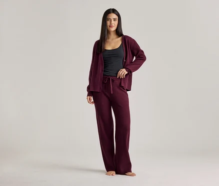 Frau in schwarzem Top, weinroter Strickjacke und weinroter Hose. Sie steht barfuß vor einem hellen Hintergrund.