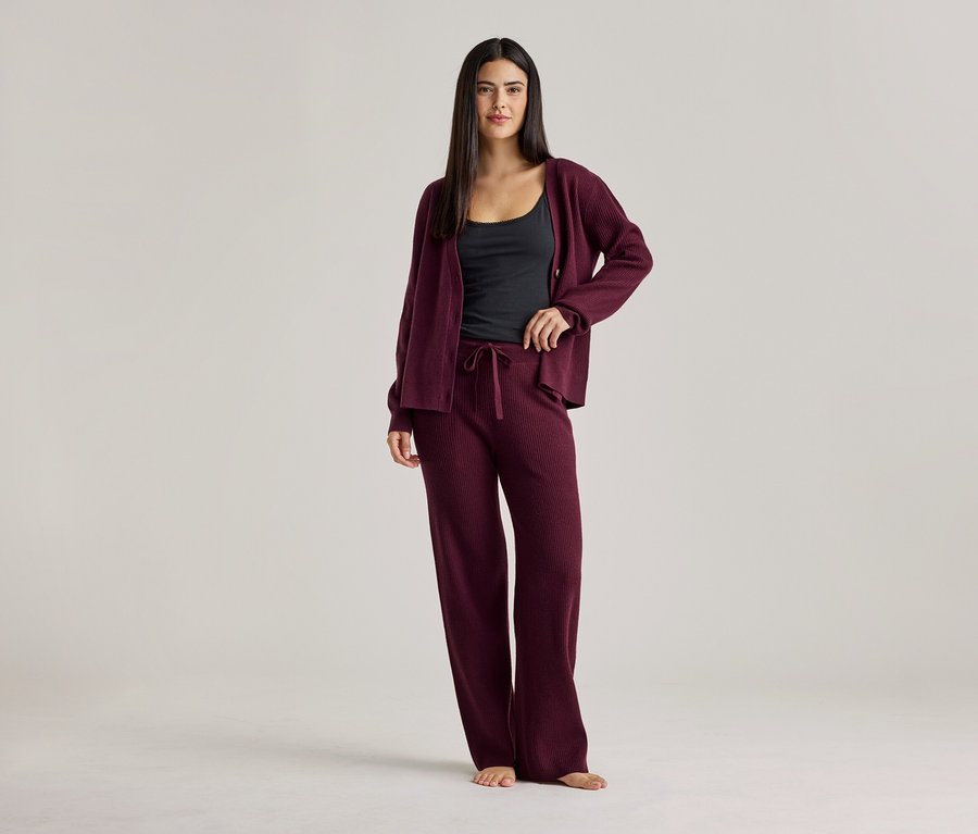 Frau in schwarzem Top, weinroter Strickjacke und weinroter Hose. Sie steht barfuß vor einem hellen Hintergrund.