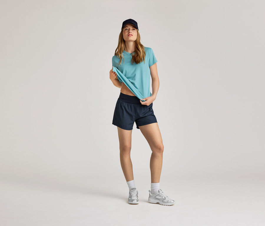 Frau mit marineblauer Kappe, türkisfarbenem T-Shirt und marineblauen Shorts. Sie trägt weiße Socken und silberne Turnschuhe.