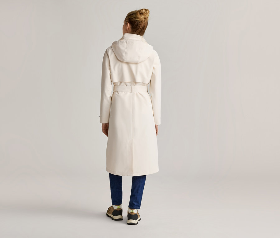 Rückansicht einer Frau mit Dutt, die einen langen, hellen Trenchcoat mit Kapuze, Gürtel und blaue Jeans mit Turnschuhen trägt.