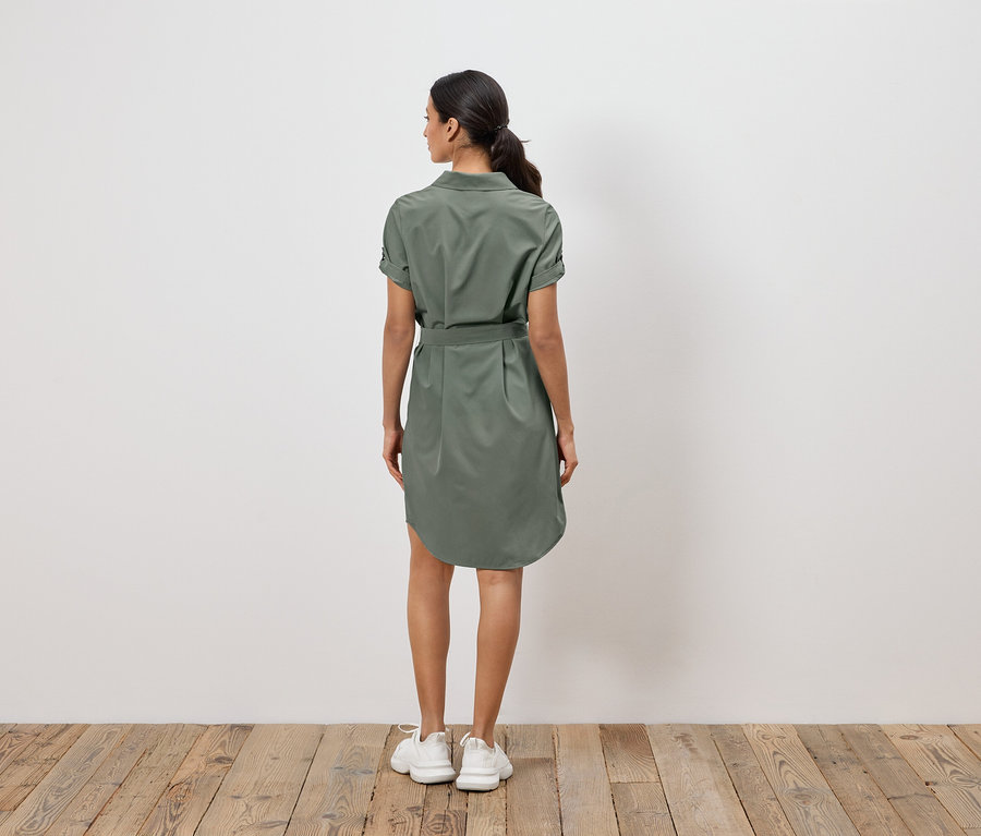 Eine Frau steht mit dem Rücken zur Kamera und trägt ein olivgrünes Hemdkleid mit Gürtel und weiße Turnschuhe.