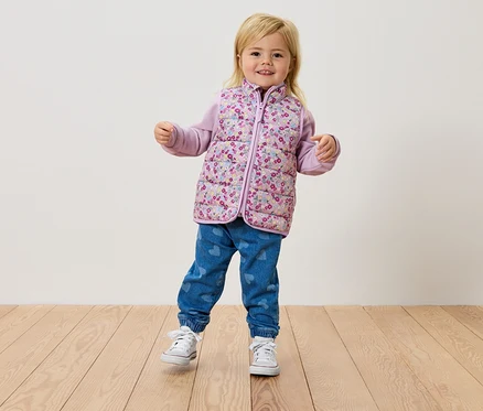Ein kleines blondes Mädchen steht auf einem Holzboden und trägt eine Blumenweste, ein lila Oberteil und Jeans.
