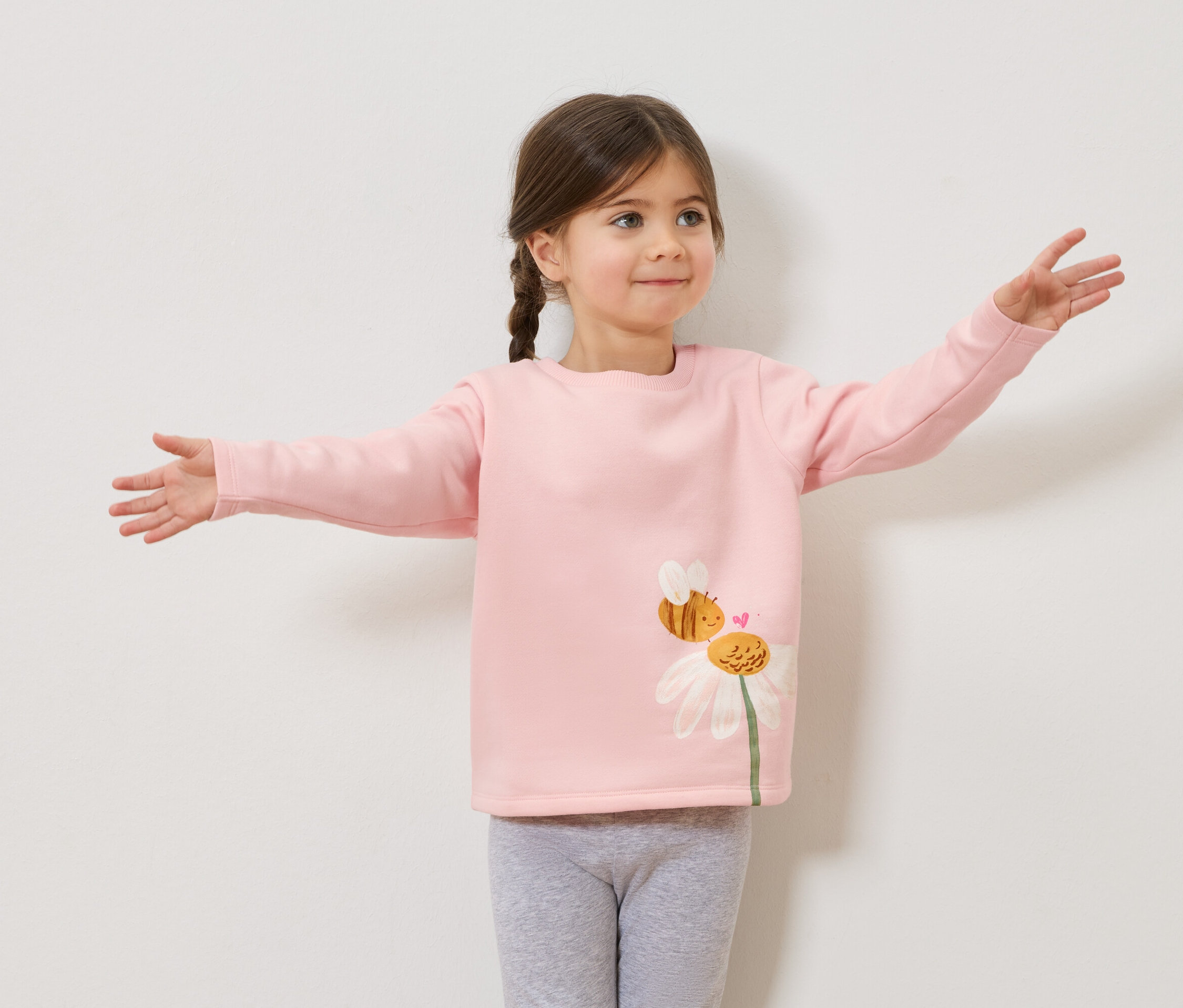 Mädchen mit Zöpfen und rosa Pullover mit Bienen- und Gänseblümchenaufdruck, graue Leggings, Arme ausgestreckt.