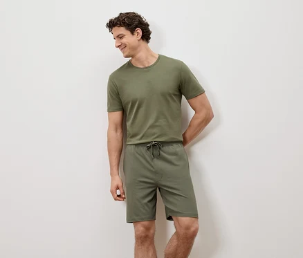 Ein Mann lehnt an einer weißen Wand und trägt ein grünes T-Shirt und Shorts.