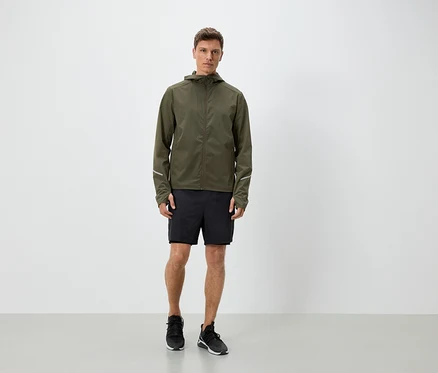 Mann in olivgrüner Jacke mit Kapuze, schwarzen Shorts und Turnschuhen.