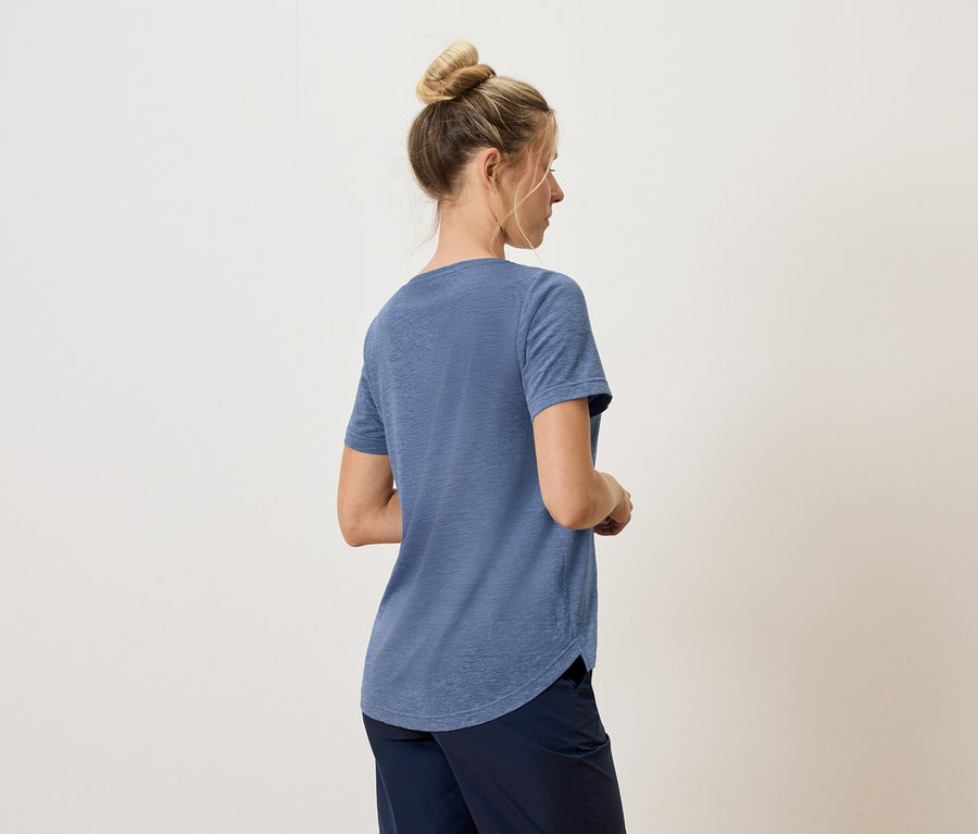 Frau von hinten, trägt blaues T-Shirt und dunkle Hose vor neutralem Hintergrund.