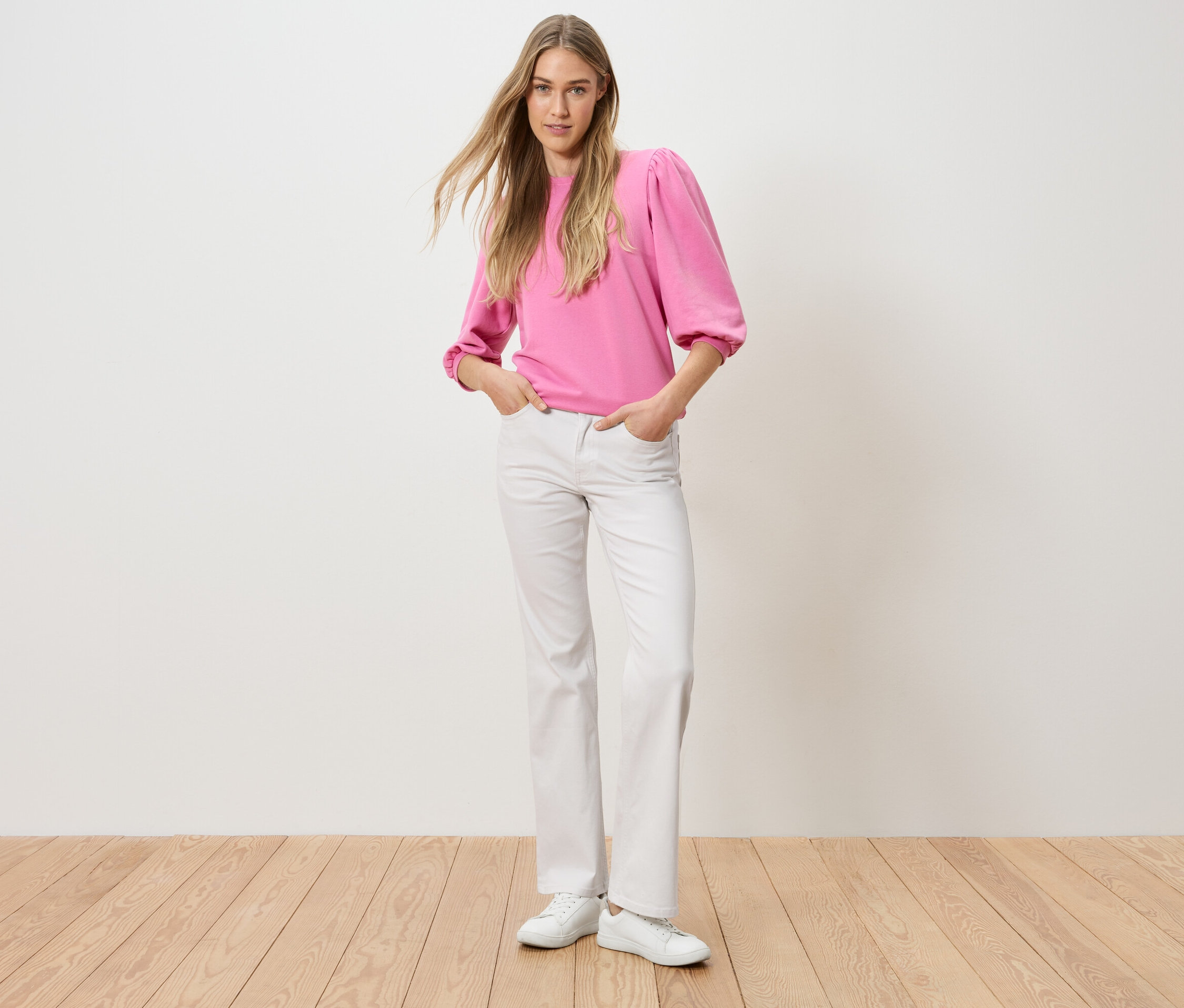 Eine Frau steht mit ihren Händen in den Hosentaschen. Sie trägt ein pinkes Oberteil und weiße Jeans.