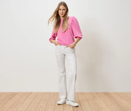 Eine Frau steht mit ihren Händen in den Hosentaschen. Sie trägt ein pinkes Oberteil und weiße Jeans.
