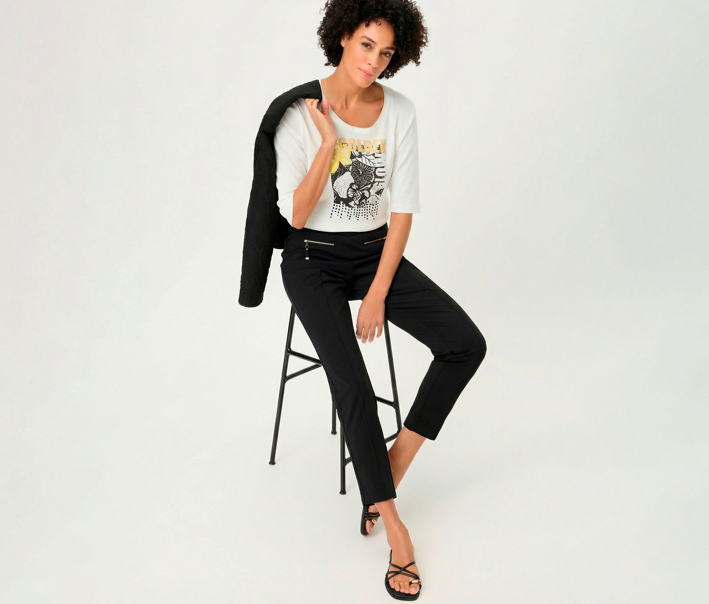 Frau sitzt mit Olsen 3/4-Shirt auf einem Hocker, Jacke über der Schulter.