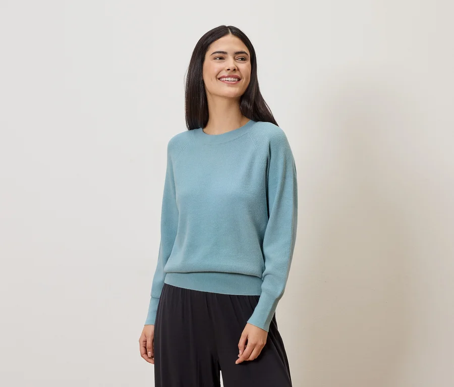Lächelnde Frau mit langen, dunklen Haaren trägt einen hellblauen Pullover und eine weite, schwarze Hose vor einem hellen Hintergrund.