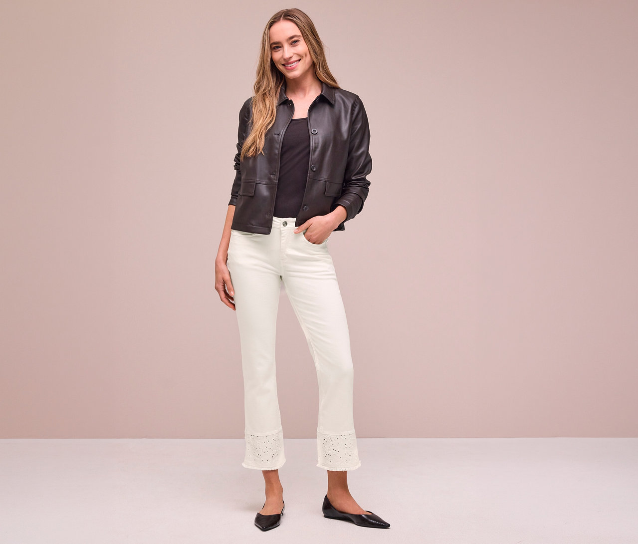 Lächelnde Frau in einer dunkelbraunen Jacke, schwarzem Oberteil und hellen Street One Jeans mit Lochstickerei am Saum, eine Hand in der Tasche.