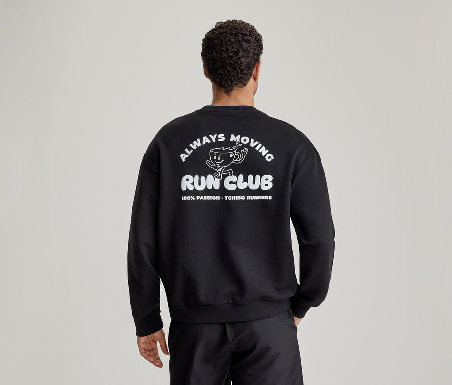 Mann von hinten in schwarzem Sweatshirt mit weißem Aufdruck 'ALWAYS MOVING RUN CLUB' und laufendem Kaffeebecher-Cartoon am Rücken.