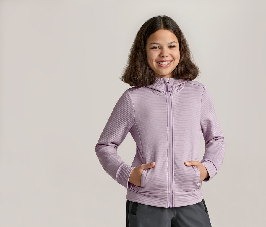 Lächelndes Mädchen mit den Händen in den Taschen einer lila Kapuzenjacke mit Reißverschluss und grauer Hose.