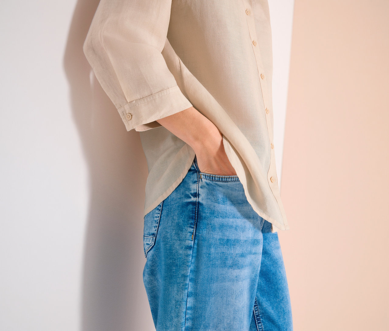 Person mit beiger Leinenbluse und blauen Jeans, Hand in der Hosentasche.