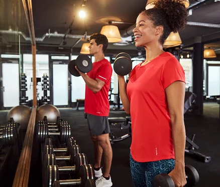 Eine Frau und ein Mann im Fitnessstudio heben Hanteln, beide tragen rote T-Shirts.